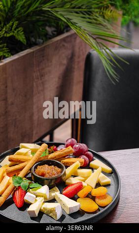 Un plateau magnifiquement agencé présente divers fromages, raisins, fraises et noix. Profiter de cette collation savoureuse tout en étant assis dans un café avec de la verdure en arrière-plan crée une atmosphère détendue. Banque D'Images