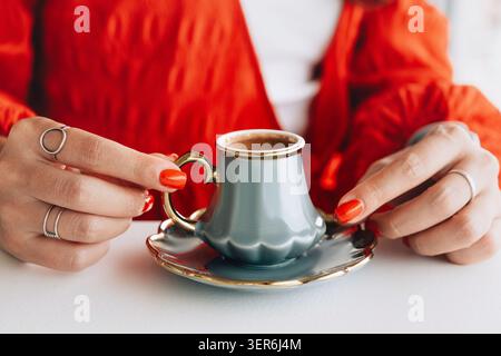 Femme dans une blouse rouge avec des ongles peints tenant une tasse de café turque traditionnelle et soucoupe, prenant une pause détente Banque D'Images