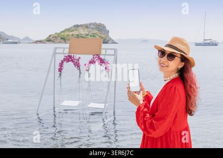 Femme souriante en robe rouge et chapeau de paille tenant un smartphone avec un écran blanc vierge contre une balançoire de l'eau de mer Banque D'Images