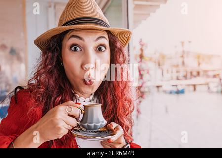 Jeune femme dans un chapeau de paille profitant des vacances, faisant un drôle de visage étonné tout en dégustant du café turc traditionnel Banque D'Images