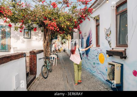 13 mai 2026, Sigacik, Turkiye : jeune femme explorant une charmante ruelle pavée avec des bougainvilliers colorés, de l'art de rue et des bâtiments blancs dans un me Banque D'Images