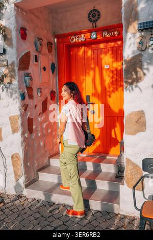 13 mai 2026, Sigacik, Turkiye : femme aux cheveux roux souriants, montant les escaliers jusqu'à une porte orange sur un mur texturé décoré de pots colorés Banque D'Images