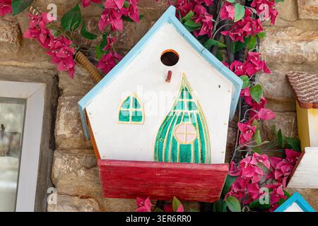 Decorative birdhouse featuring fairy tale style windows, a perch, and a flower box, set against a stone wall with vibrant pink bougainvillea Banque D'Images