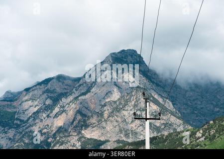 Poteau électrique et lignes électriques à haute tension contre un majestueux pic de montagne rocheuse sous les nuages bas, concept d'infrastructure régionale. Haut-v Banque D'Images