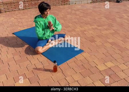 Femme assise en position de lotus avec les mains ensemble, pratiquant la pleine conscience et la concentration sur un tapis de yoga à l'extérieur Banque D'Images