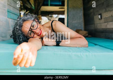 Femme souriante avec des cheveux gris bouclés et des lunettes se trouve sur un lit de camping-car, atteignant la caméra, profitant d'un voyage de vacances détendu à l'intérieur d'un va confortable Banque D'Images