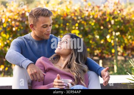Couple assis sur le banc dans le jardin ensoleillé tenant une tasse blanche portant des chandails en tricot près de la plante Banque D'Images