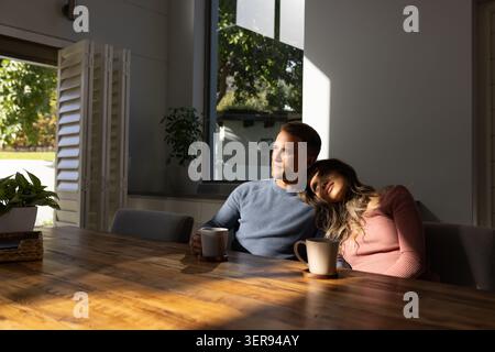 Couple diversifié assis à la table à manger en bois près de la fenêtre, appuyé sur l'épaule et tenant des tasses Banque D'Images