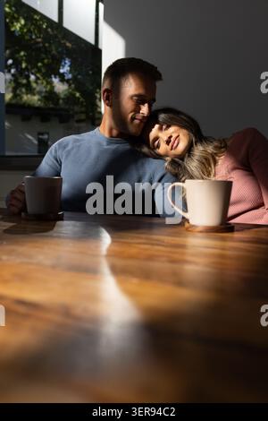 Couple diversifié portant des vêtements en tricot, assis à une table à manger en bois, penché, tenant des tasses en céramique Banque D'Images