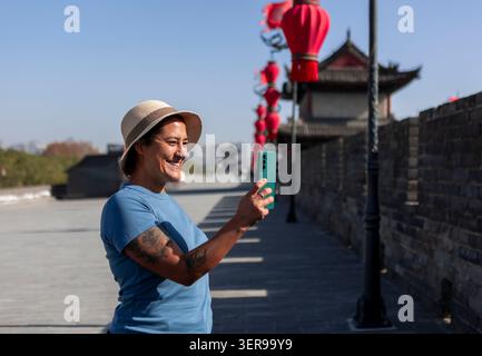 La femme souriante touriste utilise un smartphone pour prendre une photo en marchant sur l'ancien mur de la ville à Xi'an, en Chine, avec des lanternes rouges et des tours historiques Banque D'Images