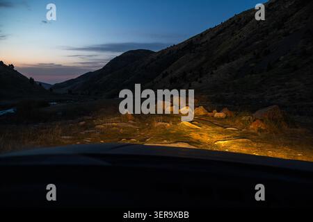 La scène crépusculaire capture la vallée de montagne vue de l'intérieur d'une voiture. Les phares éclairent un terrain rocheux et un ruisseau sinueux, créant une atmosphère sereine Banque D'Images