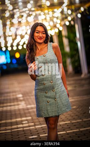 Jeune femme souriante dans une robe sans manches posant sur une passerelle de la ville la nuit, avec des cordes chaudes et un bokeh doux créant un urbain élégant et joyeux Banque D'Images