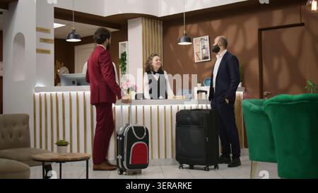 Hommes d'affaires demandant des informations sur les installations de l'hôtel, discutant avec l'employé de la réception et attendant dans le hall, avec l'équipe de direction approchant la réception pour l'enregistrement avant la prochaine conférence internationale. Banque D'Images