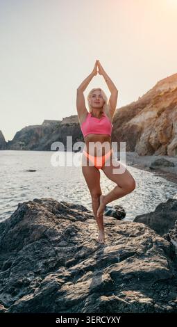 Yoga plage de bien-être femme blonde en bikini rose effectue la pose d'arbre sur les rochers côtiers au bord de la mer pour la remise en forme Banque D'Images