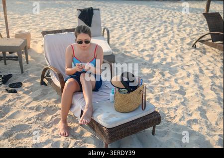 Une femme asiatique dans les années 30 est assise sur une chaise de plage, portant des lunettes de soleil et un bikini coloré. Elle est concentrée sur son téléphone, entourée de sable et de plage A. Banque D'Images