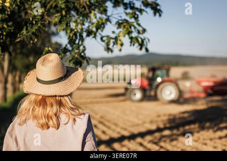 Agricultrice avec chapeau de paille regardant le tracteur travaillant sur le champ agricole à la ferme Banque D'Images