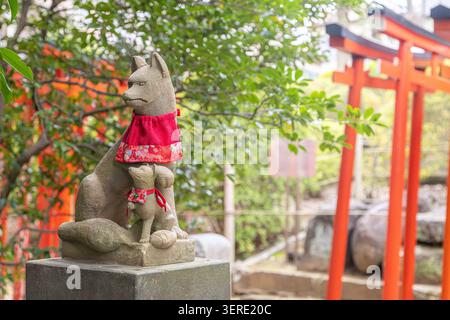 Gardien renard Inari en pierre portant un bavoir rouge à côté des portes torii vermillon au sanctuaire Otome Inari, Tokyo, Japon. Banque D'Images