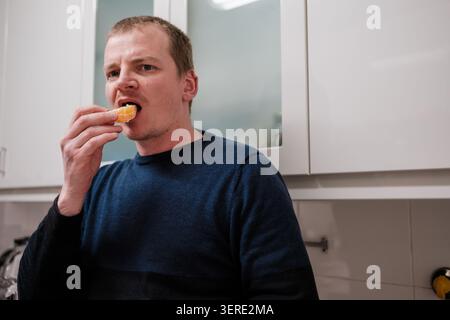 Homme appréciant une collation saine d'orange mandarine dans un cadre de cuisine moderne Banque D'Images