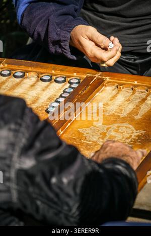Gros plan de deux mains d'hommes engagés dans une partie de backgammon sur une planche de bois bien usée. Un joueur tient un dé blanc, prêt à faire un mouvement pendant un s. Banque D'Images