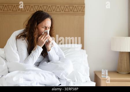Femme éternuant dans les tissus tout en se reposant dans le lit, montrant des symptômes de rhume Banque D'Images