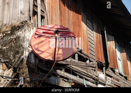 Une antenne parabolique rouge pâle sous la lumière directe du jour projette une forte ombre contre le côté d'une vieille maison en bois en ruine avec du béton altéré. Banque D'Images