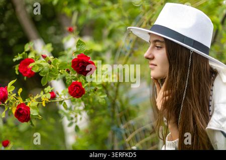 Jeune femme au chapeau blanc admirant les roses rouges. Jeune femme dans un chapeau blanc regarde des roses rouges en fleurs dans un jardin vert luxuriant. Banque D'Images