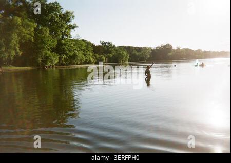 Une personne se tient à la taille dans un lac serein avec le bras levé, entouré d'arbres verdoyants et de lumière dorée du soleil, évoquant des vibrations estivales insouciantes. Banque D'Images