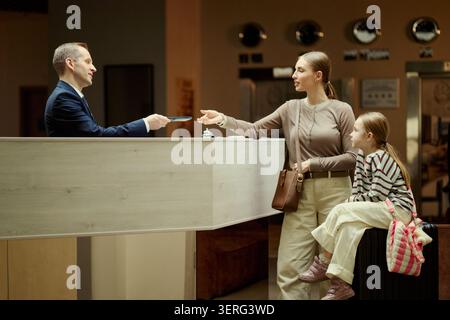 Grand plan d'une mère caucasienne arrivant à la réception de l'hôtel avec enfant et bagages, recevant la carte-clé de la réceptionniste. Idéal pour la réservation de voyage, tourisme familial, annonces hôtelières Banque D'Images