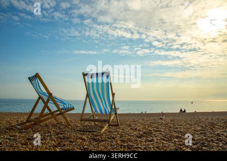Deux chaises longues rayées sur la plage de Brighton au soleil du début de soirée d'automne avec des silhouettes lointaines au bord de l'eau Banque D'Images