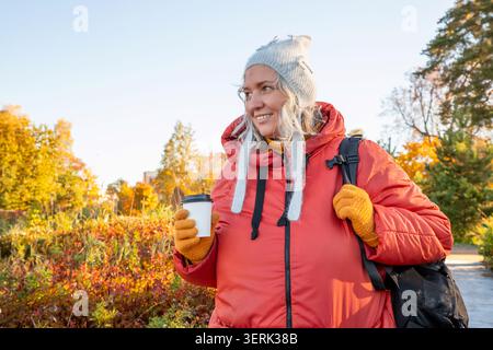 Femme d'âge moyen appréciant le café à emporter sur la promenade d'automne dans le parc de la ville. Voyageur portant une veste rouge bouffante et bonnet tricoté chaud tenant la tasse. Santé l Banque D'Images