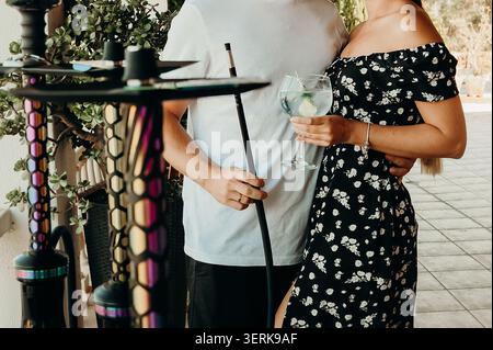 Couple apprécie les boissons et narguilé dans un cadre extérieur lumineux avec des plantes lors d'un week-end de rassemblement. l'homme et la femme se tiennent proches l'un de l'autre, appréciant le drin Banque D'Images