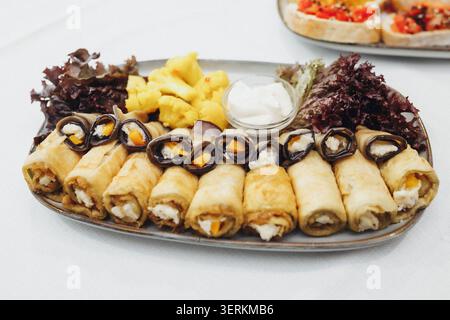 Rouleaux dorés remplis de fromage crémeux et de fruits. Des morceaux d'ananas jaune vif sont assis à côté d'eux. Les légumes verts frais et un petit bol de trempette ajoutent de la couleur. S Banque D'Images