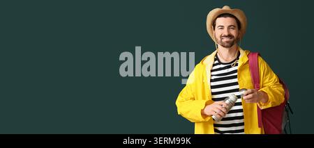 Jeune homme souriant en imperméable jaune et chapeau de paille tenant thermos avec sac à dos, prêt pour l'aventure en plein air Banque D'Images