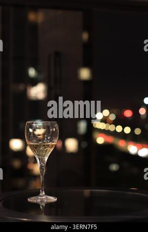 Un verre à vin contenant une petite quantité de vin blanc sur une table réfléchissante, photographié la nuit avec des lumières de ville colorées et un flou artistique doux en arrière-plan Banque D'Images