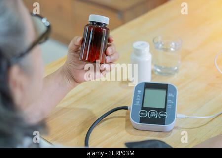 Vue arrière d'une femme âgée tenant une bouteille de pilule tout en utilisant un dispositif médical pour vérifier sa mesure de pression artérielle. Souffrant d'hypertension. Banque D'Images