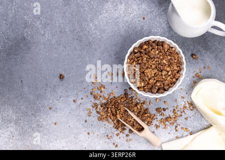 Céréales granola dans un bol blanc avec une cuillère en bois et de l'avoine dispersée sur un fond gris, prêt à ajouter le lait du pichet blanc Banque D'Images