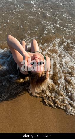 Femme en maillot de bain noir couché sur la plage de sable, éclaboussant ludique de l'eau avec les mains, la lumière du soleil se reflétant sur les vagues de l'océan, scène de loisirs d'été Banque D'Images