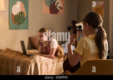 Plan moyen d'une écolière pratiquant le violoncelle tandis qu'un autre enfant étudie la musique sur ordinateur portable à la maison, soutenant l'éducation musicale à distance et le marketing de leçons en ligne pour les plates-formes d'apprentissage Banque D'Images