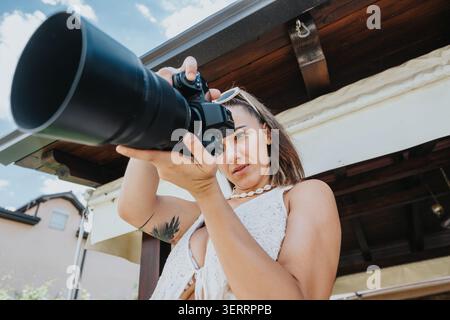 Femme photographe utilisant un appareil photo reflex numérique avec un grand objectif à l'extérieur d'un patio en bois Banque D'Images