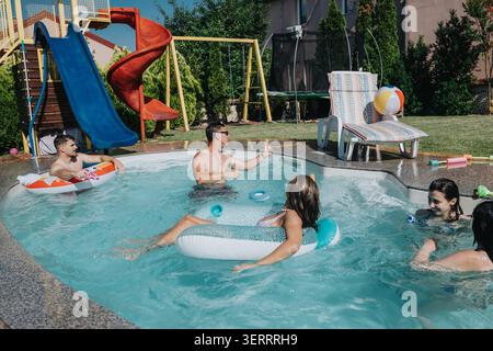 Amis profitant d'une piscine d'été dans la cour arrière avec flotteurs et toboggan sur une journée ensoleillée Banque D'Images
