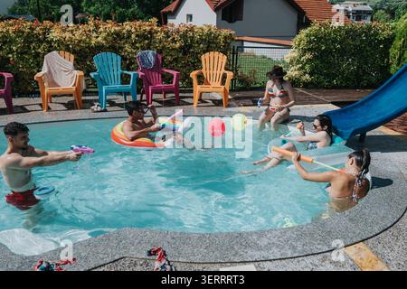 Amis profitant d'une journée d'été à la piscine avec flotteurs, jouets aquatiques et chaises de piscine détendues Banque D'Images