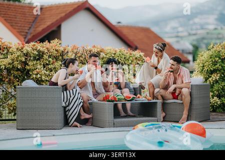 Amis appréciant l'été au bord de la piscine manger de la pastèque et se détendre sur le mobilier de patio extérieur Banque D'Images