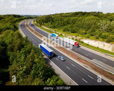 Vue aérienne du paysage d'une autoroute britannique très fréquentée avec des camions et des voitures coupant une bande à travers la campagne boisée reliant les grandes villes sur un su lumineux Banque D'Images