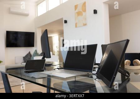 Ordinateur portable argenté assis ouvert sur le bureau en verre dans le bureau à domicile ouvert, casque beige à proximité Banque D'Images