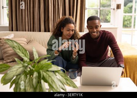 Couple assis sur un canapé beige penché vers un ordinateur portable argenté, femme pointant le smartphone, homme souriant Banque D'Images