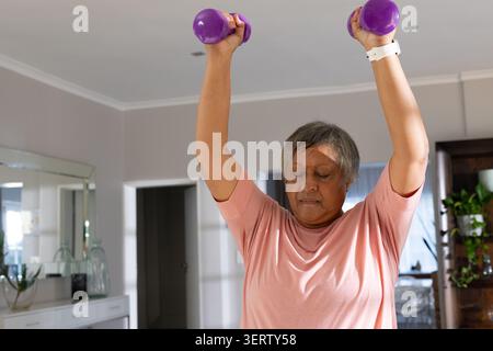 Femme africaine senior soulevant des haltères violets au-dessus de la tête dans le salon portant un tee-shirt rose montre blanche Banque D'Images