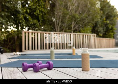 Les haltères violets reposent sur la terrasse en bois près de tapis bleus près de la piscine avec des bouteilles, balustrade en bois Banque D'Images