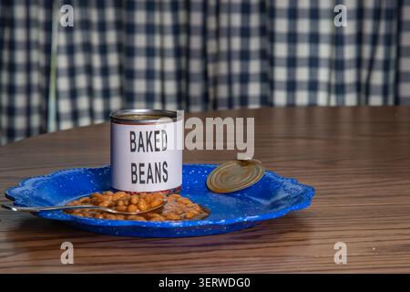 Une boîte de haricots cuits repose sur une assiette bleue à côté d'une cuillère. Les haricots sont sucrés et étalés sur l'assiette. Le cadre est une cuisine simple avec un rideau à carreaux en arrière-plan. Banque D'Images