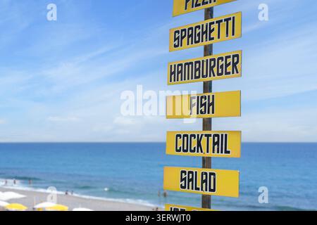Le panneau de menu jaune présente des aliments comme le poisson, cocktail, salade, avec l'océan et le ciel visibles en arrière-plan Banque D'Images