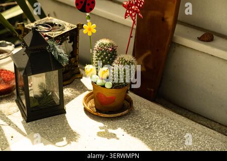 Un petit cactus mignon dans un pot en forme de coeur se trouve sur un rebord de fenêtre à côté d'une lanterne, avec quelques décorations festives suspendues à proximité. Banque D'Images
